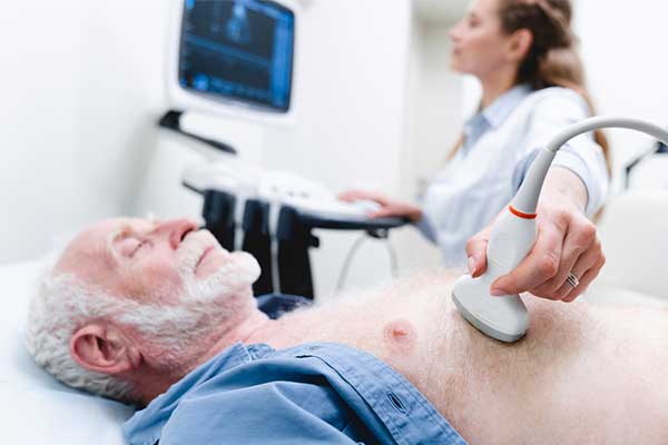 Male patient laying in a bed with his eyes closed while a medical care provider does a machine reading on his chest.